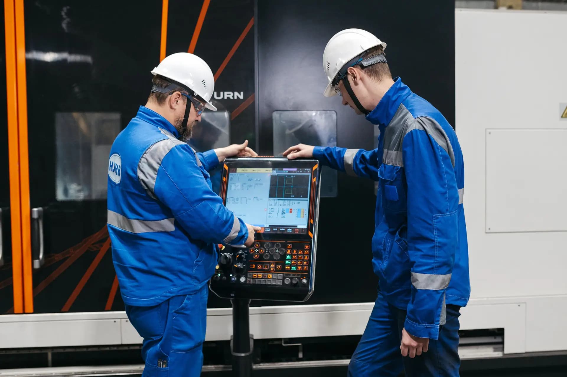 Two operators in blue workwear at a CNC machine control panel