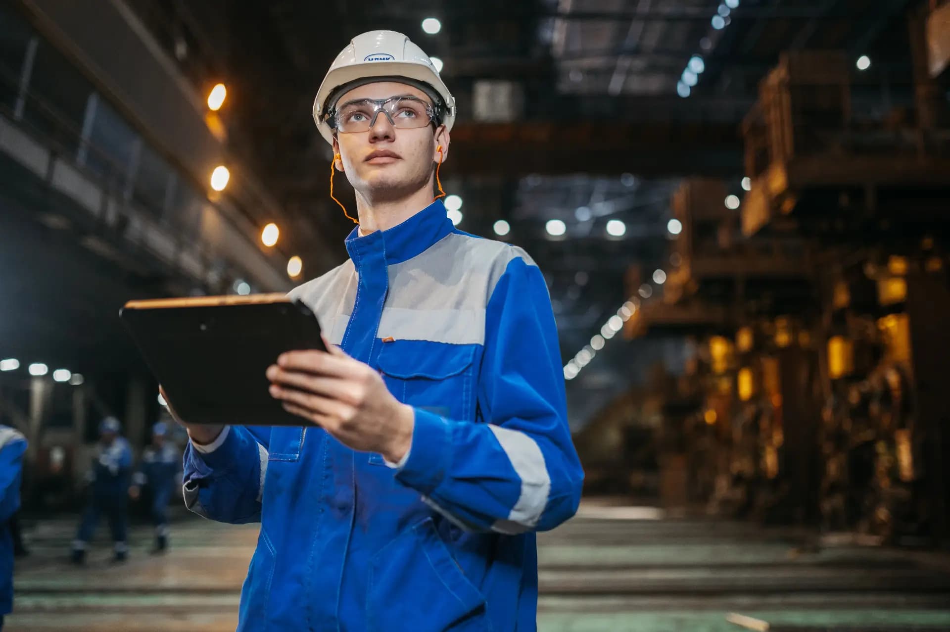 Engineer in a hard hat holding a tablet in a heavy-industry hall