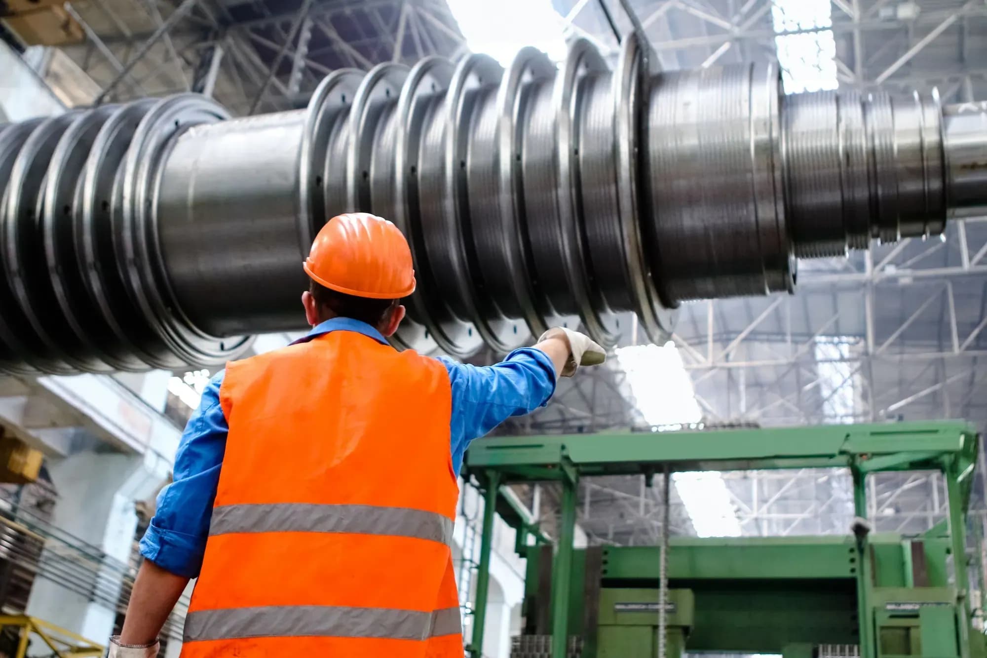 Engineer in an orange hard hat and hi-vis vest pointing at a large machine shaft in a plant hall