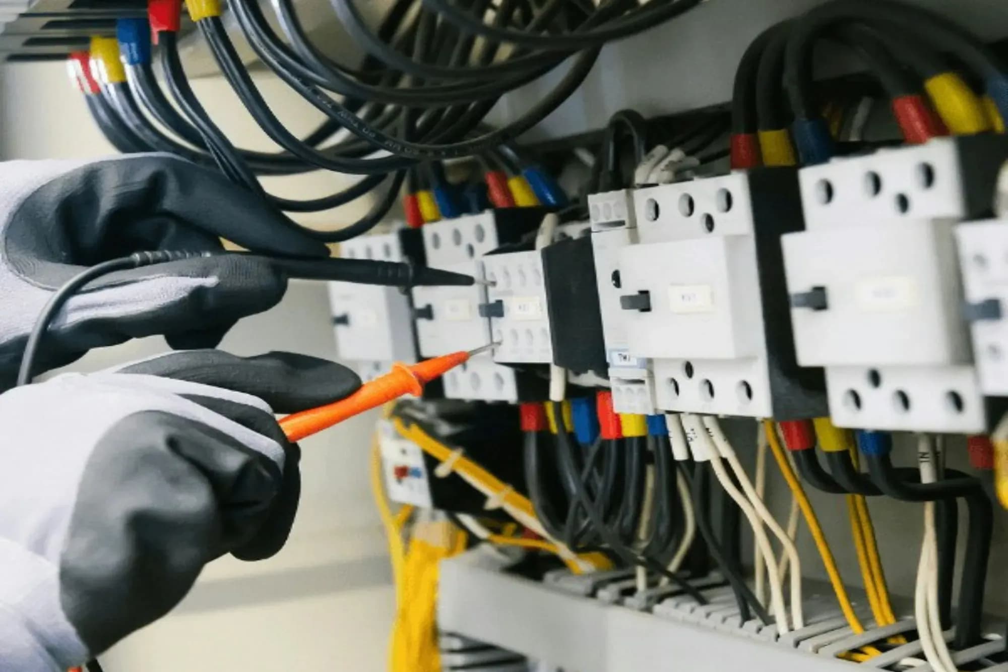 Gloved hands holding two multimeter probes against contactor terminals with coloured ferruled wires in a control panel