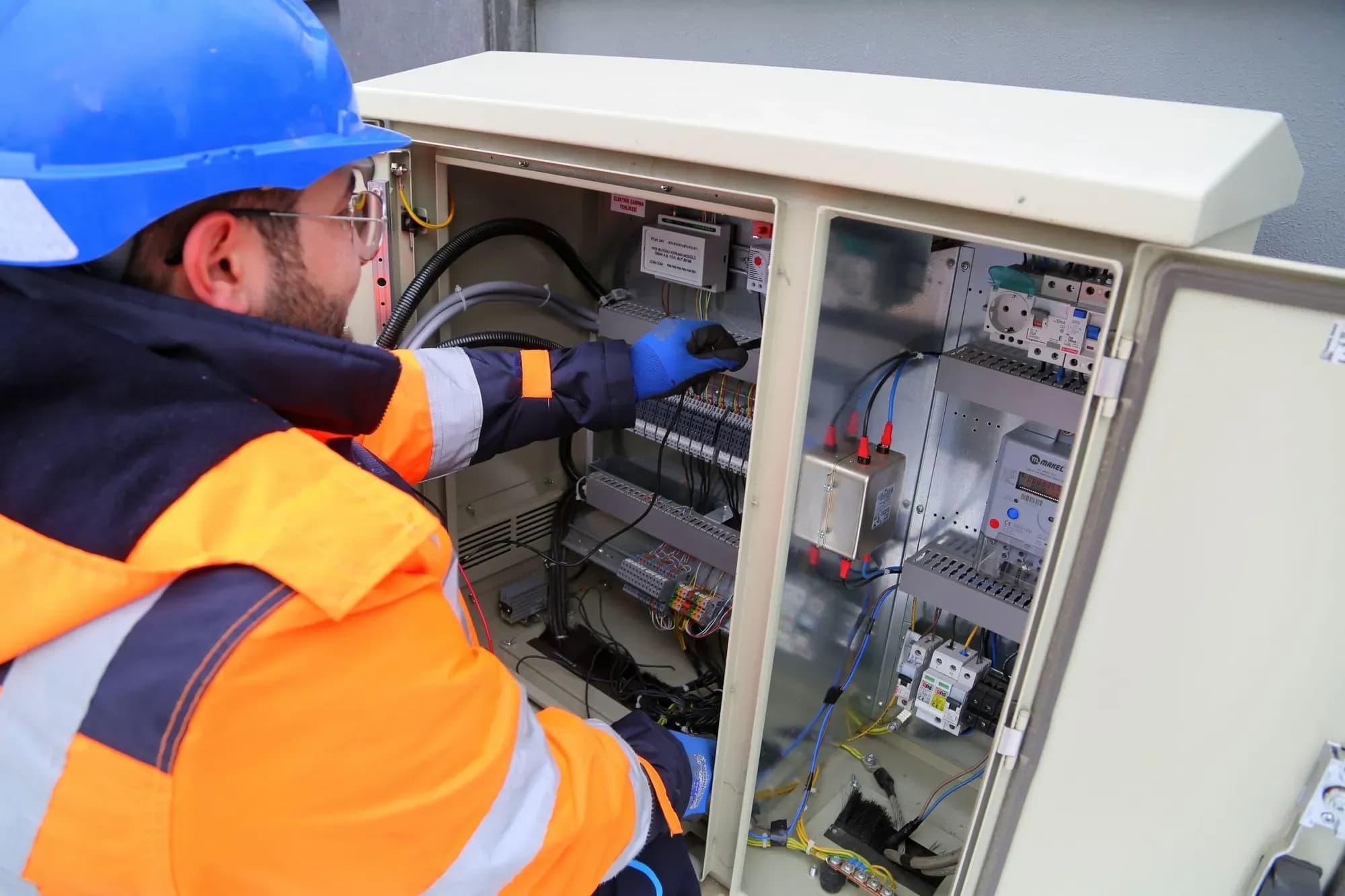 Maintenance engineer in a blue hard hat and orange hi-vis checking breakers inside an outdoor electrical cabinet