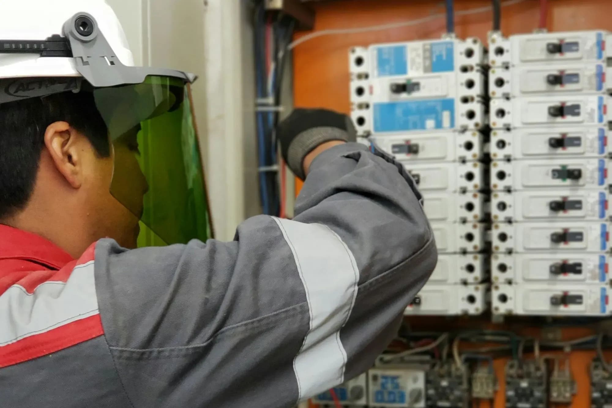 Electrician in a hard hat and face shield testing breakers in an open electrical panel with a multimeter