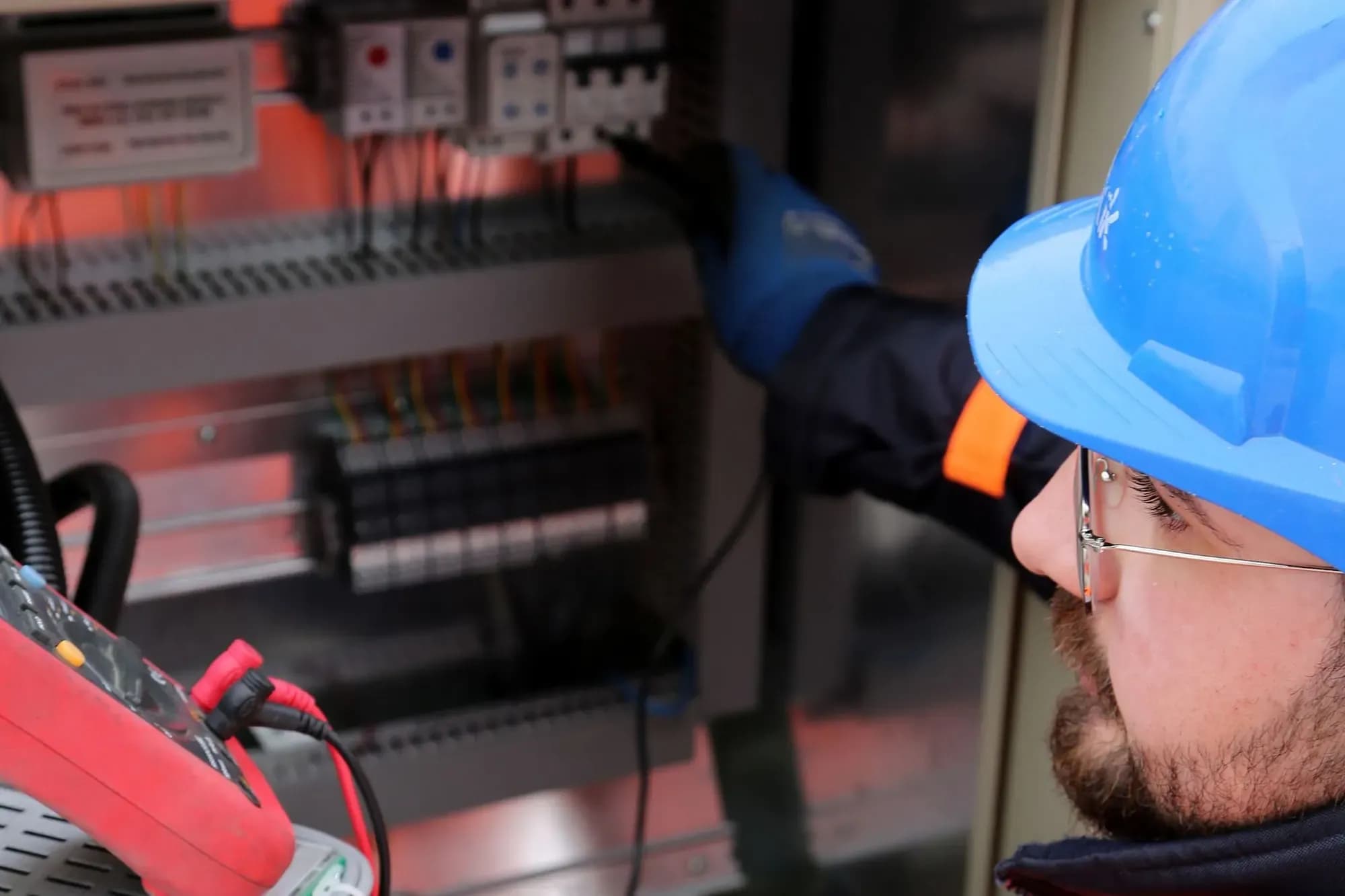 Engineer in a blue hard hat and orange hi-vis jacket measuring inside a control panel with a clamp meter