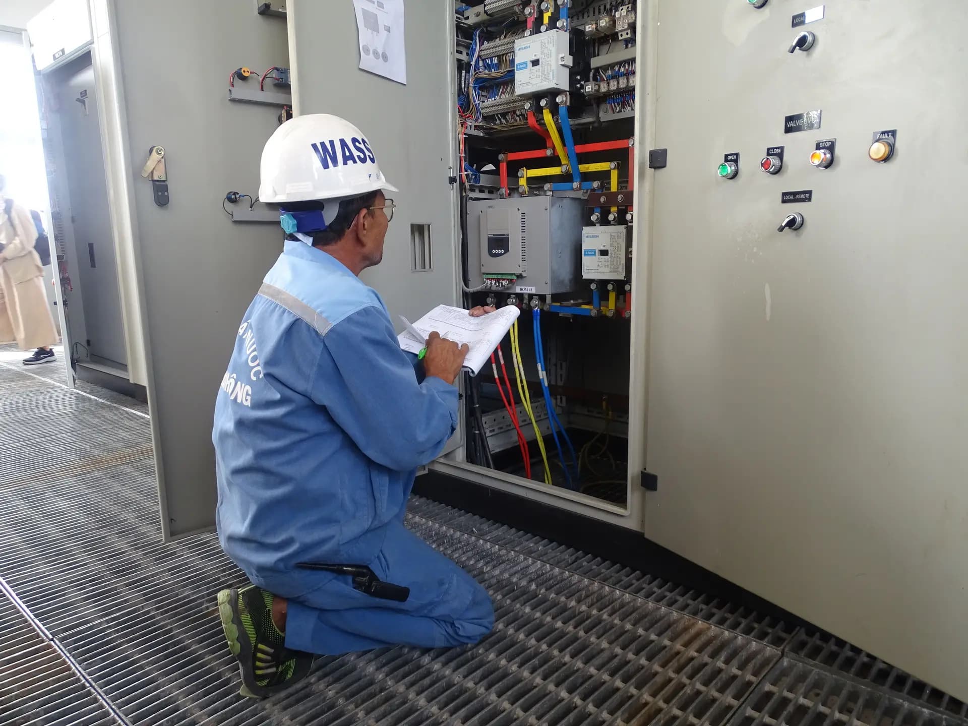 Engineer in a white hard hat checking a switchboard and noting readings on a clipboard