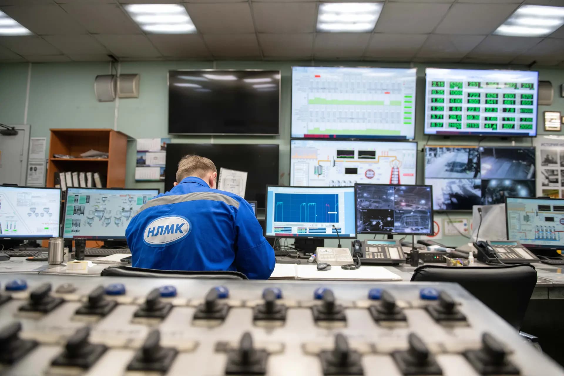 Operator at a desk in a plant control room with a wall of monitoring screens