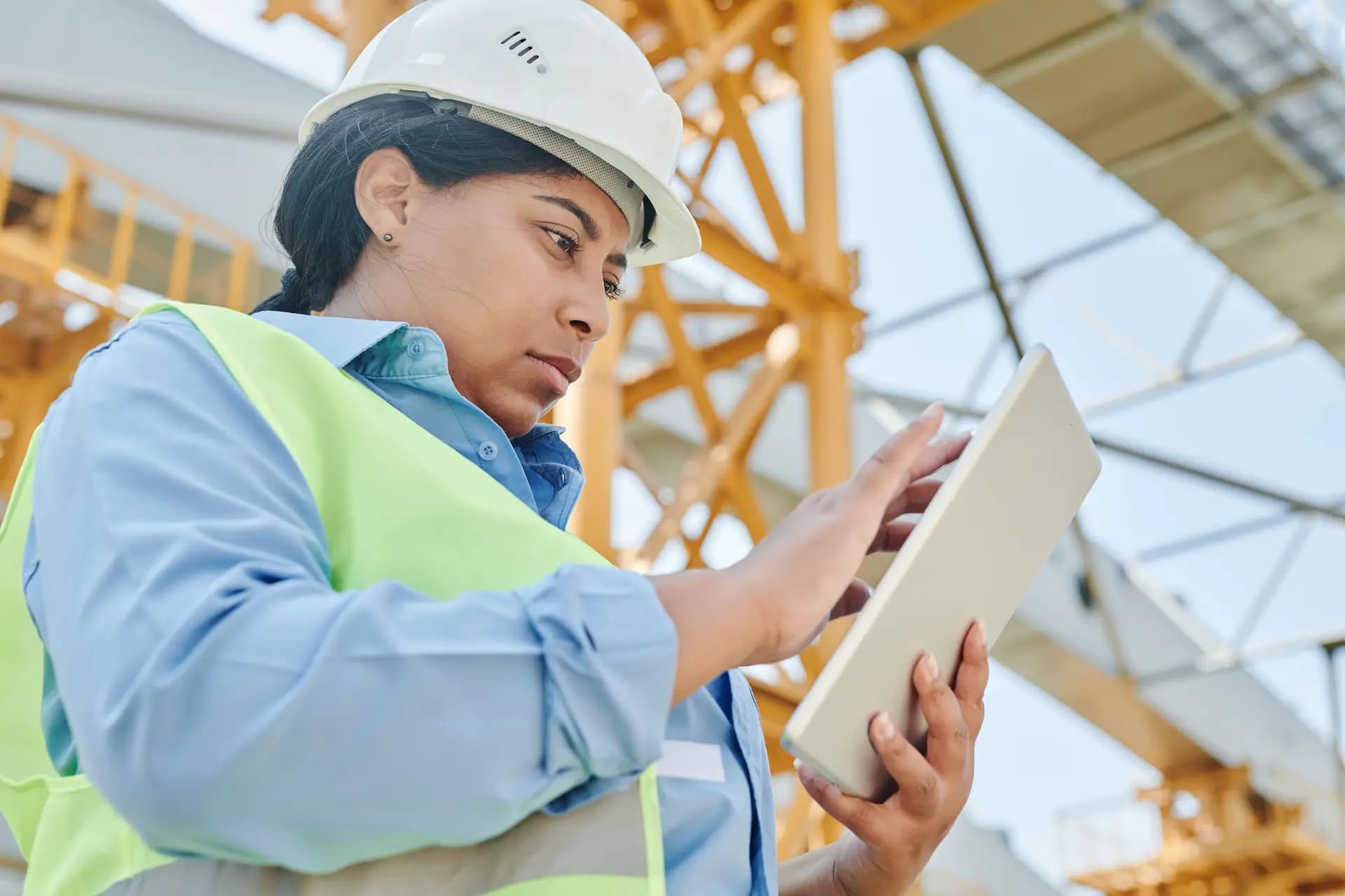Woman in a hard hat and hi-vis vest reviewing a tablet outdoors beneath a crane