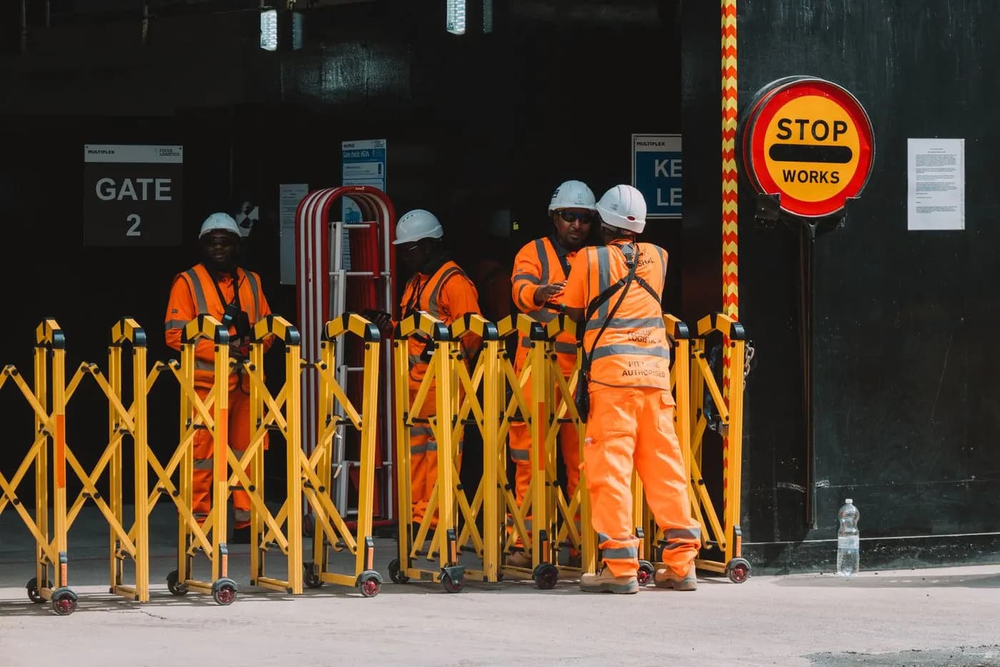 Site crew in orange hi-vis and white hard hats beside a yellow barrier and a Stop Works sign at a London construction gate