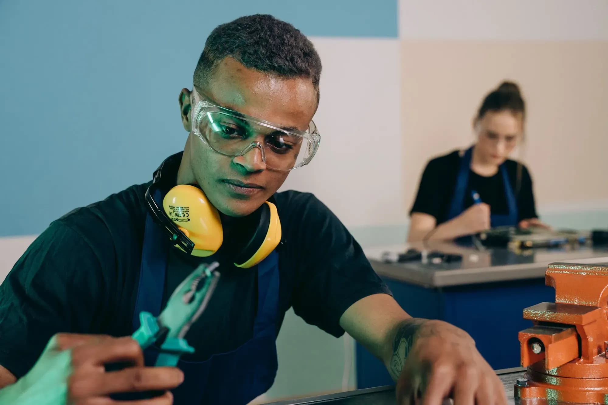 Apprentice in safety glasses and ear defenders holding pliers at a workbench, with a colleague working behind
