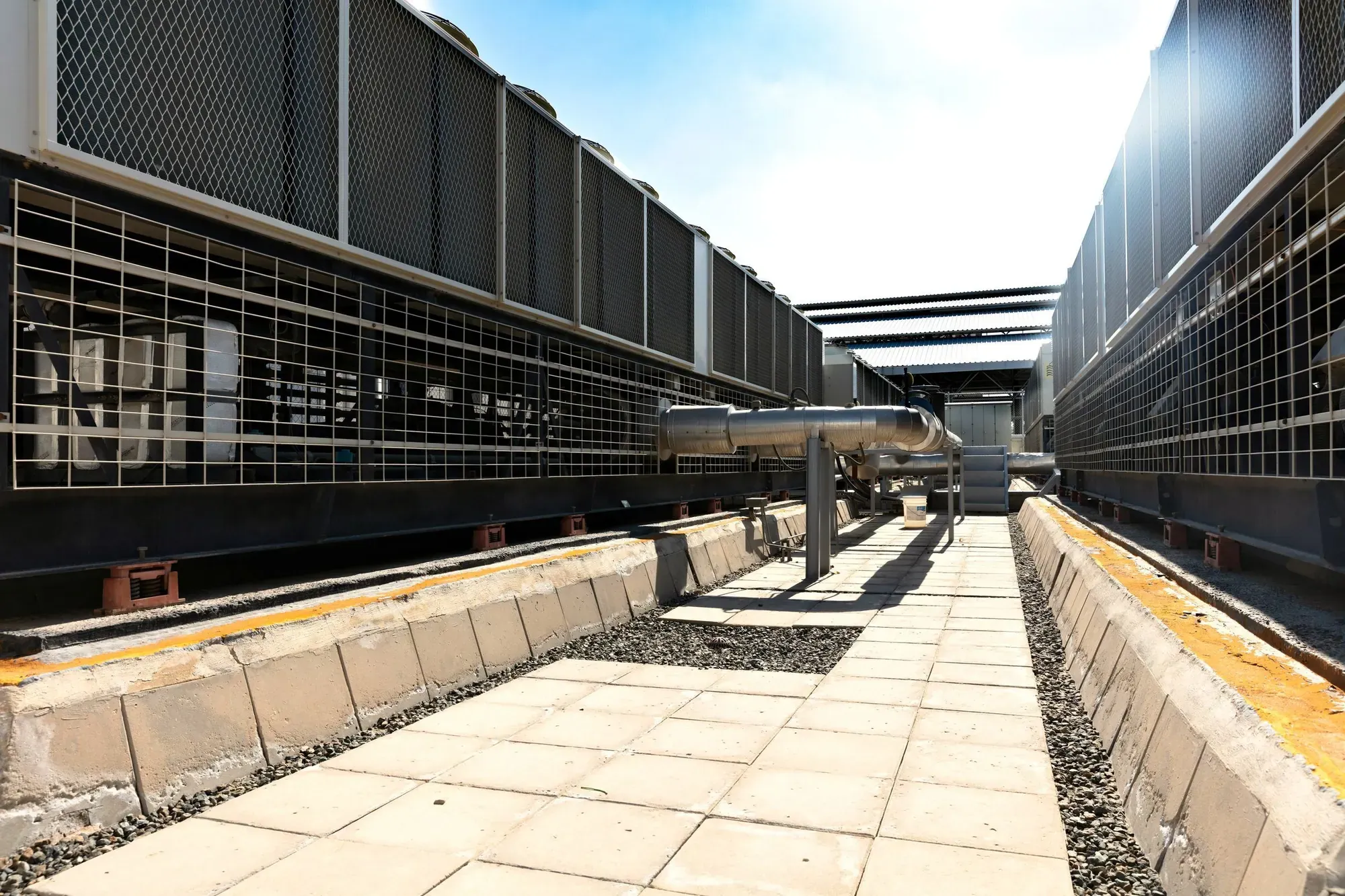 Rows of large air-cooled chiller units with metal guard grilles and insulated pipework beside a paved walkway