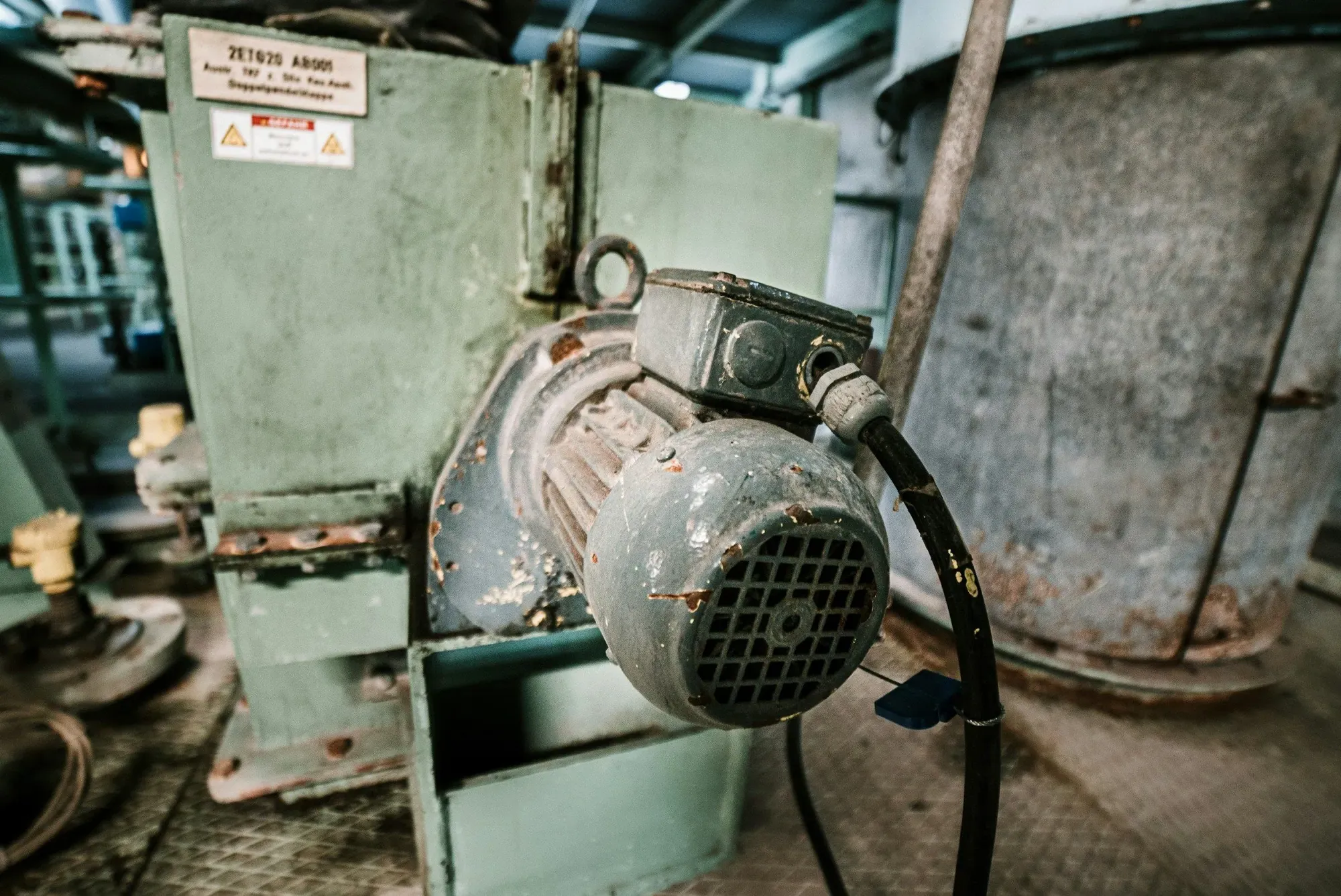 Old three-phase electric motor with a cast terminal box and cable gland mounted on green plant machinery. A motor's terminal box is where the winding ends are connected.