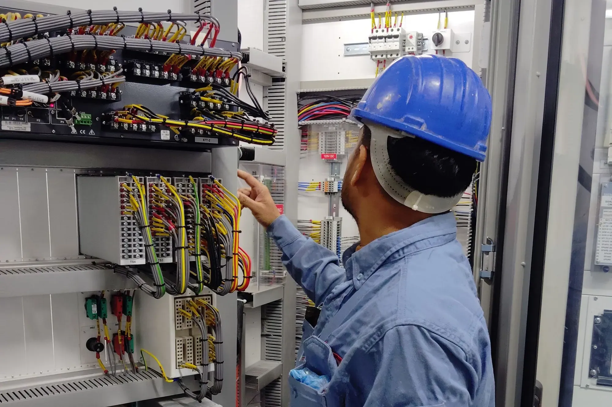 An electrician in a blue hard hat checking wiring inside a tall industrial control cabinet
