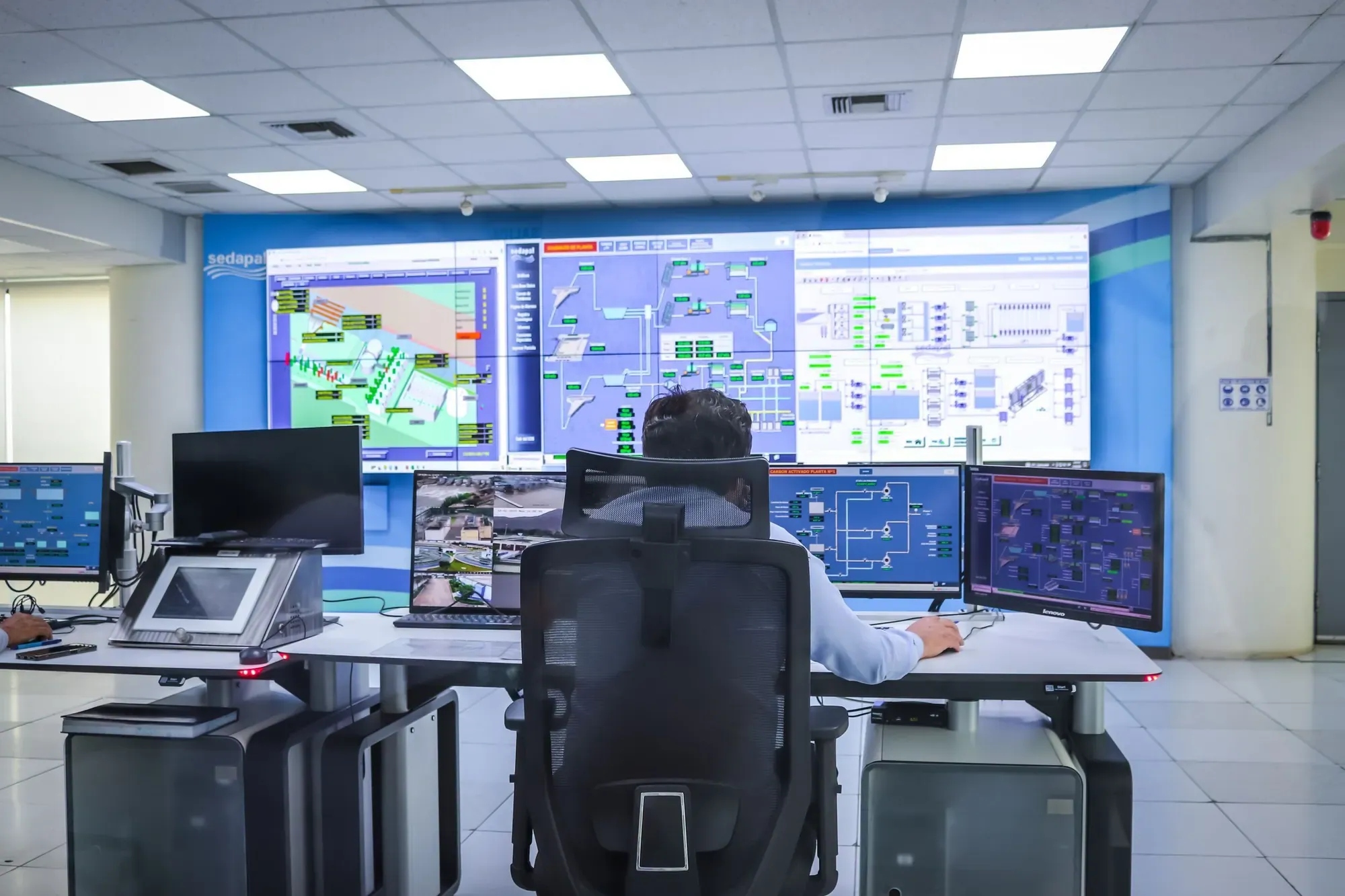 Operator at a desk in a modern control room facing a video wall of process screens, the kind of site that is staffed round the clock